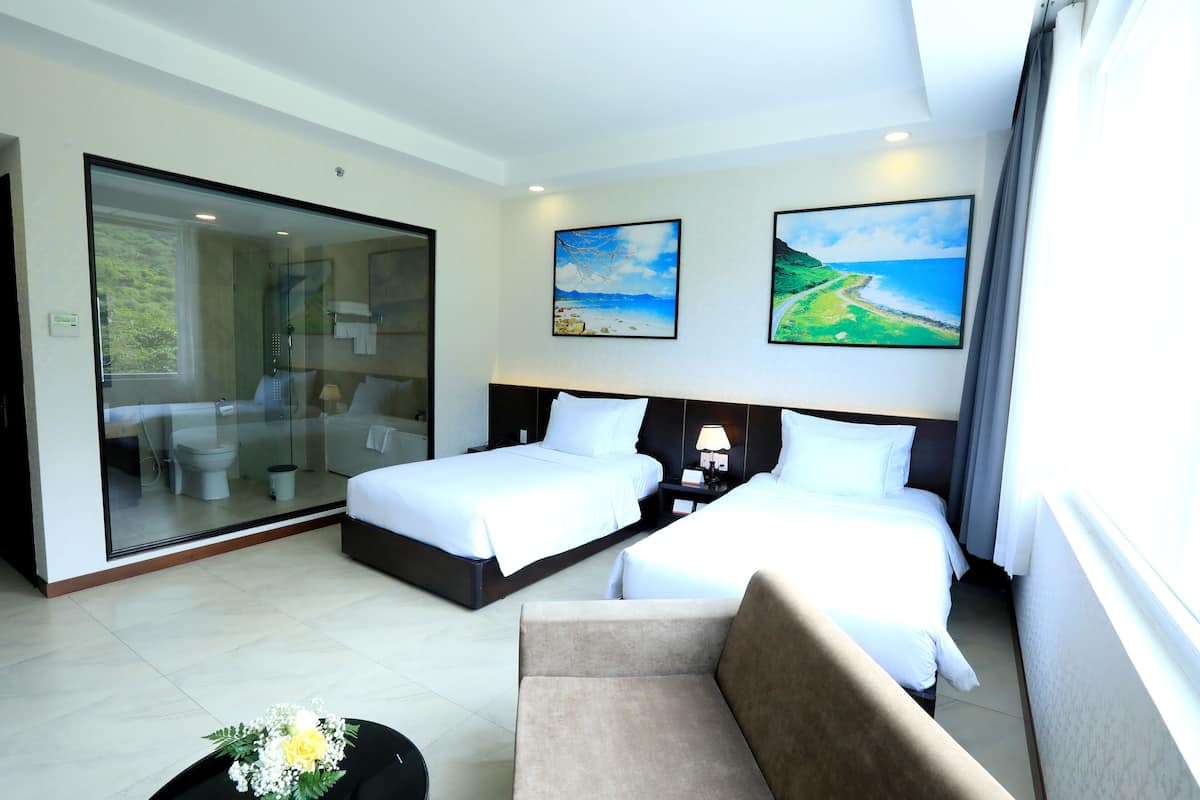 Quarto de hotel com duas camas, um sofá, área de chuveiro em vidro e duas pinturas nas paredes.