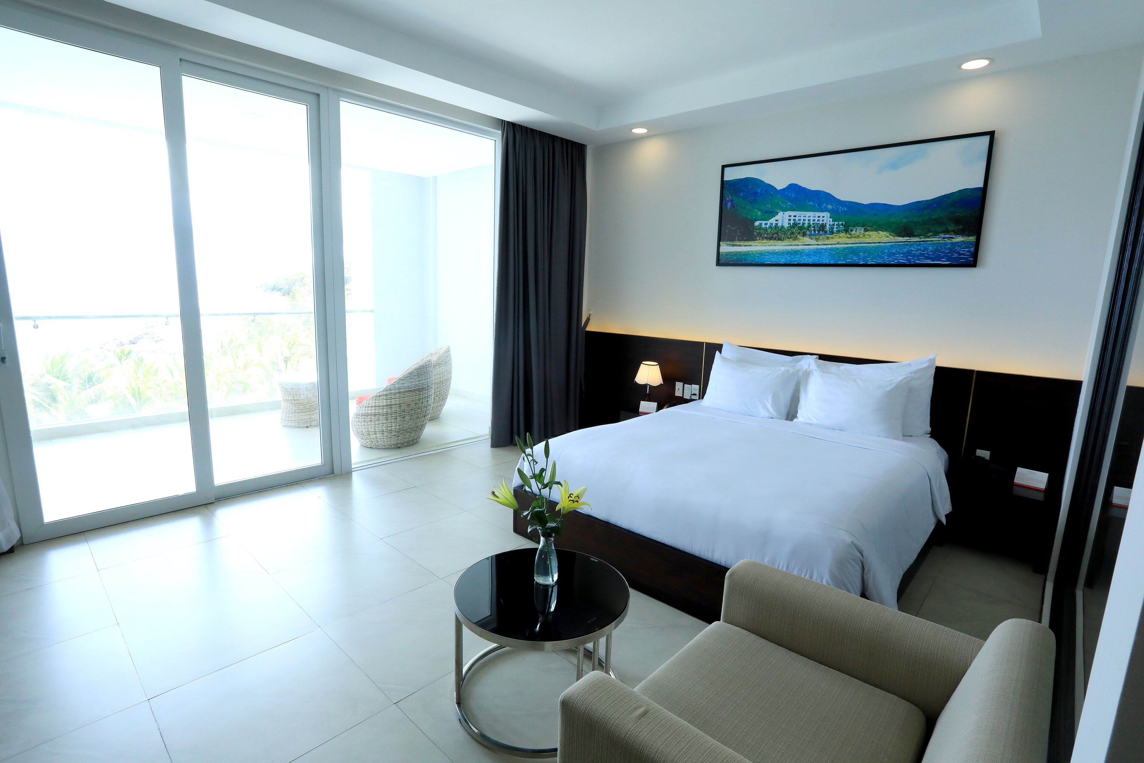 A hotel room with a large bed, a round table with a vase, a sofa, a nightstand, and a painting of a landscape on the wall.