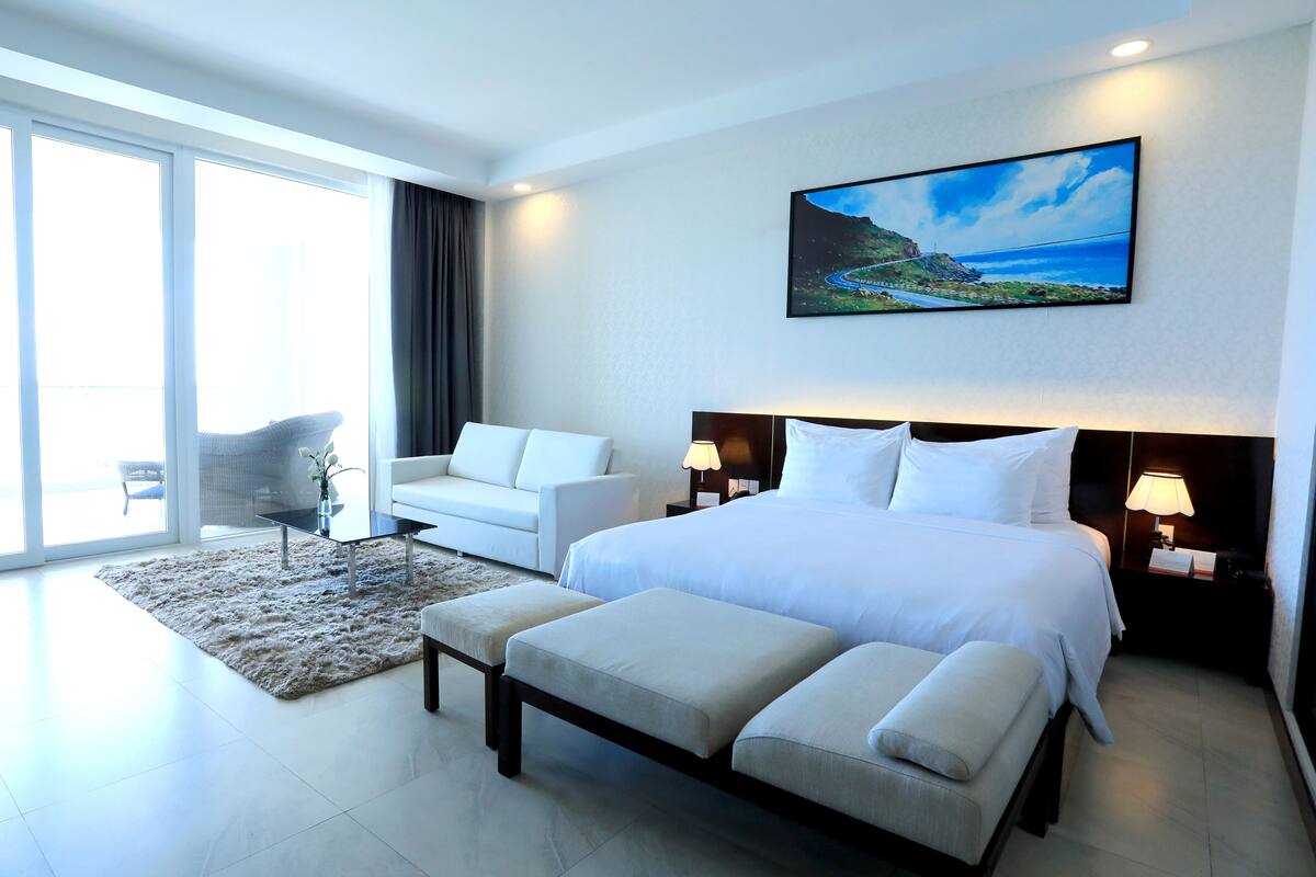 Quarto de hotel moderno com uma cama grande, um sofá, uma mesinha e vista para o exterior.