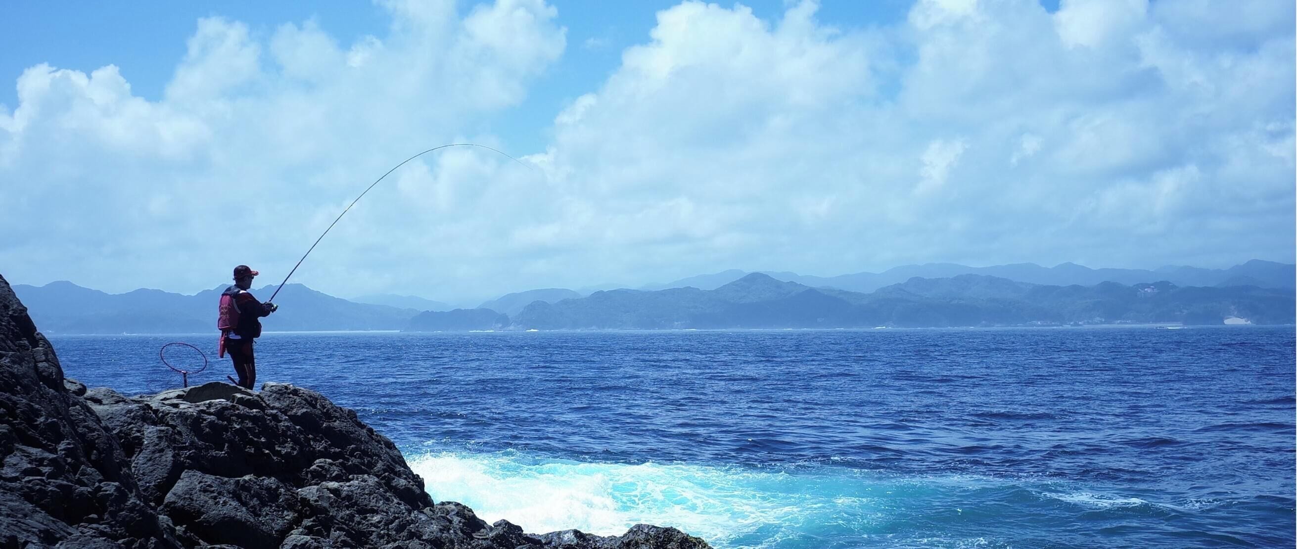 Une personne est en train de pêcher au bord d’une côte rocheuse, avec une vue sur des montagnes lointaines et l’océan.