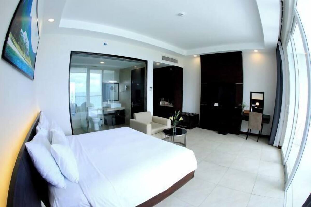 Quarto de hotel moderno com uma cama grande, área de estar com uma mesinha, escrivaninha e uma janela ampla.