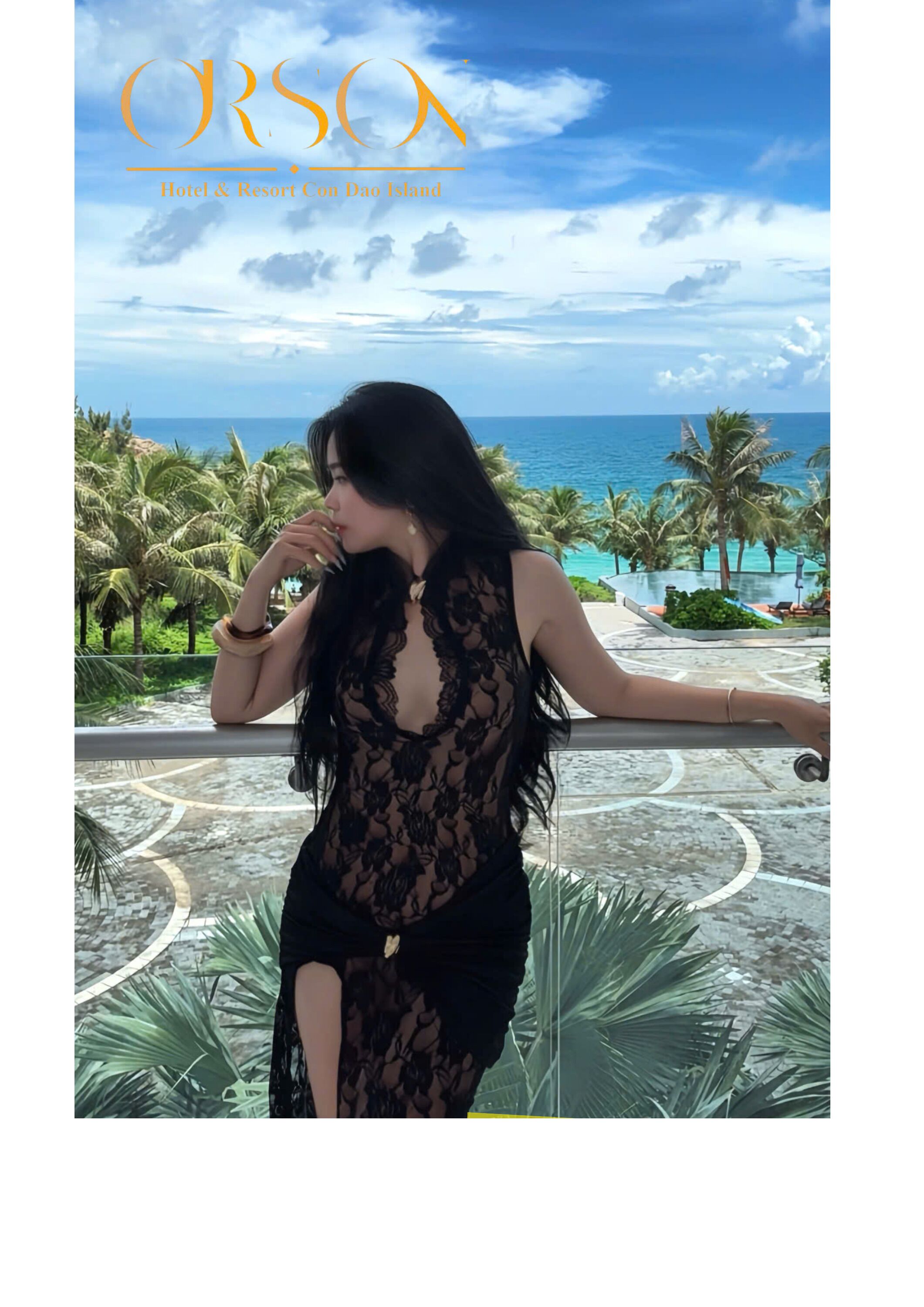 A woman in a black lace dress standing by a pool with a view of the ocean and palm trees.