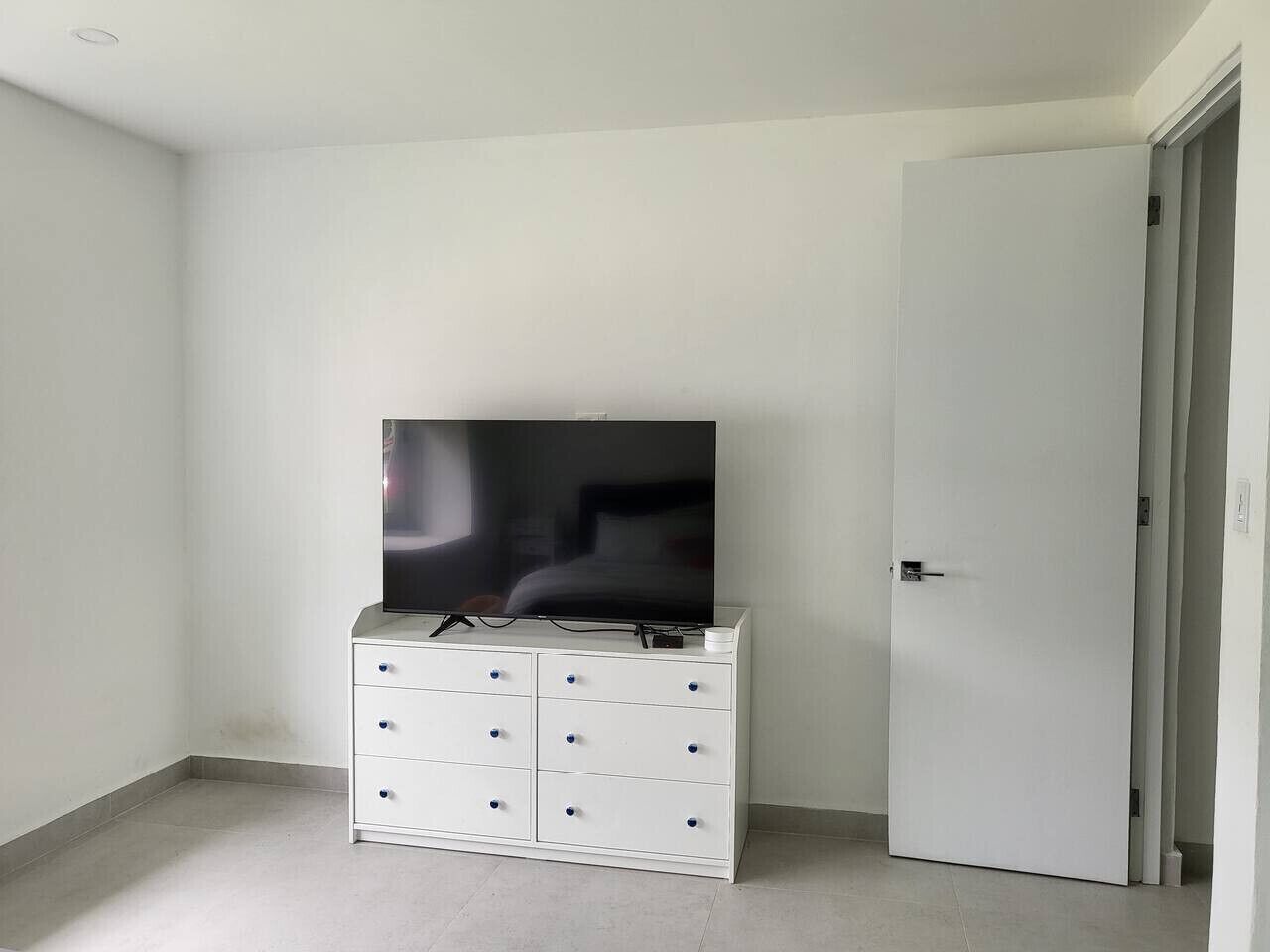 Quarto com cômoda branca, televisão de tela plana em cima e porta fechada.