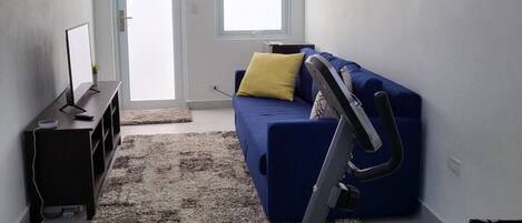 A living room with a blue sofa, a standing fan, a treadmill, and a painting on the wall.