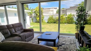 Ein Wohnzimmer mit braunem Sofa, einem schwarzen Couchtisch und Blick auf eine grüne Rasenfläche sowie weiße Gebäude durch große Fenster.