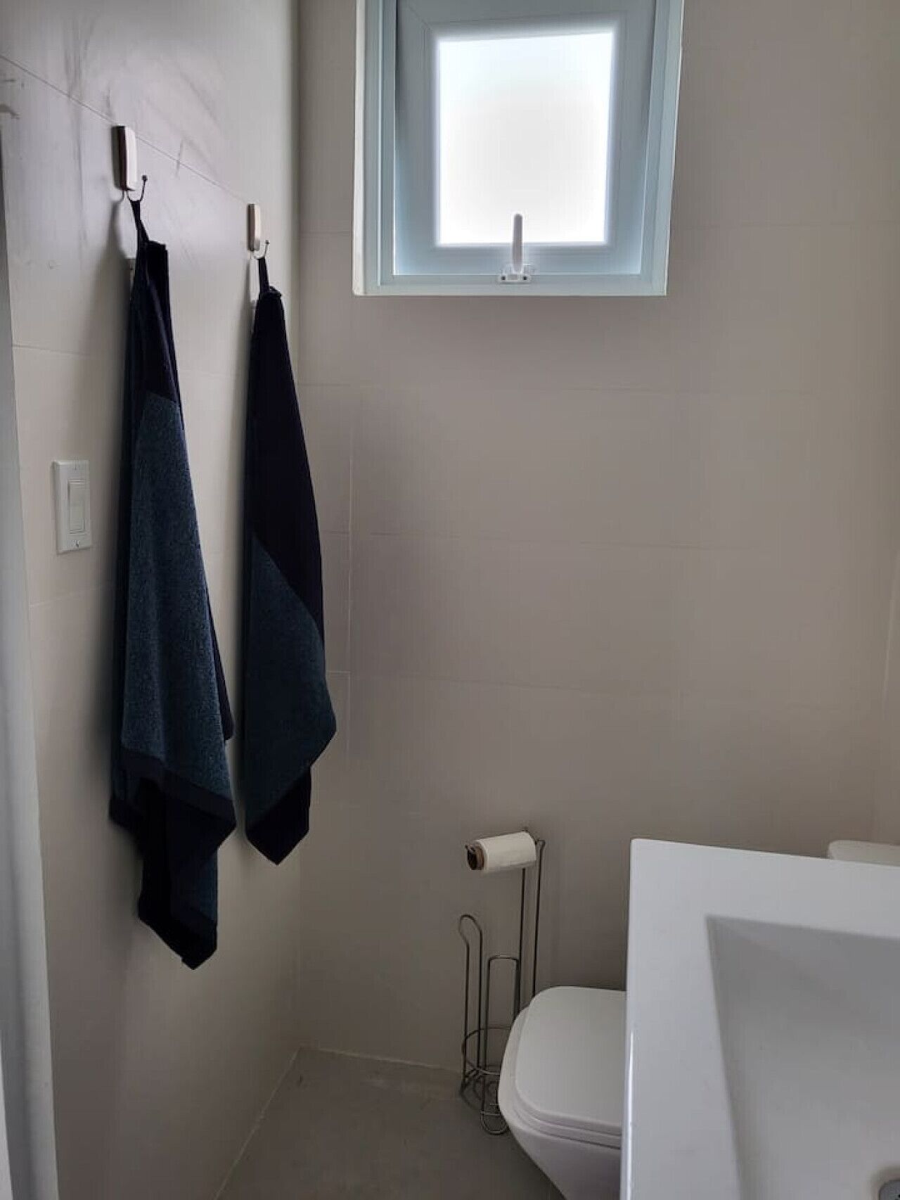 Un baño con inodoro, toallero con dos toallas, ventana y un rollo de papel higiénico.