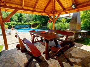Ein überdachter Essbereich im Freien mit Holzbänken und einem Tisch, ein Steine-Pool und ein Grill.