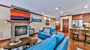 A living room with a stone fireplace, a blue sofa, a dining area with wooden chairs, and a kitchen in the background. - Prime Walk-to-Heavenly Townhome w/ A/C | TW702 (South Lake Tahoe)