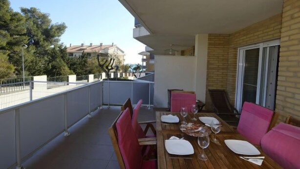 A wooden table set for dining on a balcony with pink chairs, a glass of wine, and a plate. - Residencial Mar I by Interhome (Alcanar)