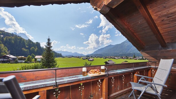 Ein Balkon mit Holzgeländer, ein Tisch mit einer Speiseplatte und zwei Bierkrügen, mit Blick auf eine grüne Landschaft und Berge im Hintergrund.