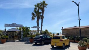 Un parking où se trouvent un taxi jaune, une voiture de couleur sombre et un bâtiment sur lequel on peut voir une enseigne indiquant la présence d’un restaurant et d’une pizzeria.
