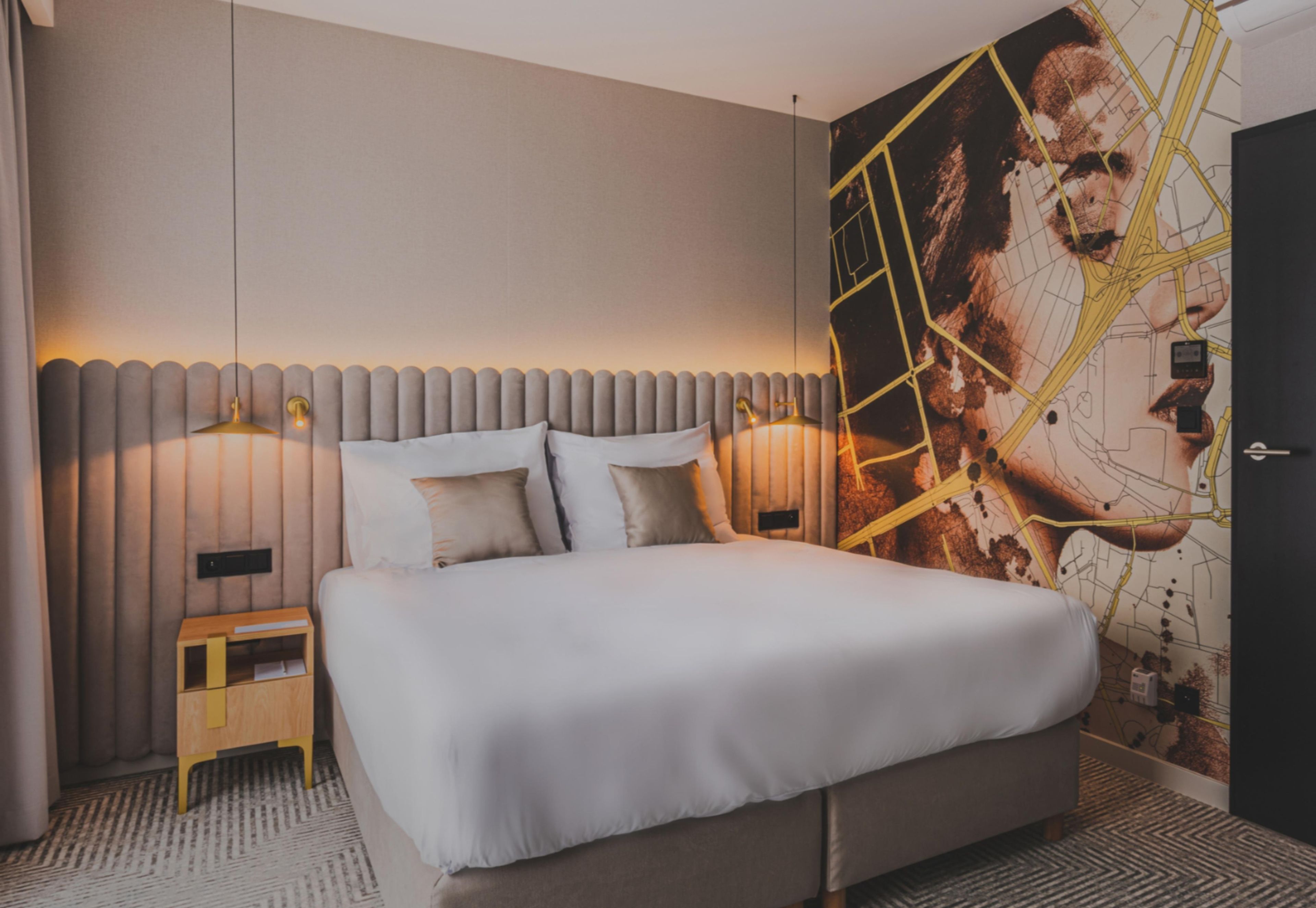 Une chambre d’hôtel moderne avec un grand lit, deux lampes de chevet, une petite table de nuit en bois et un mur orné d’une carte et d’œuvres d’art.