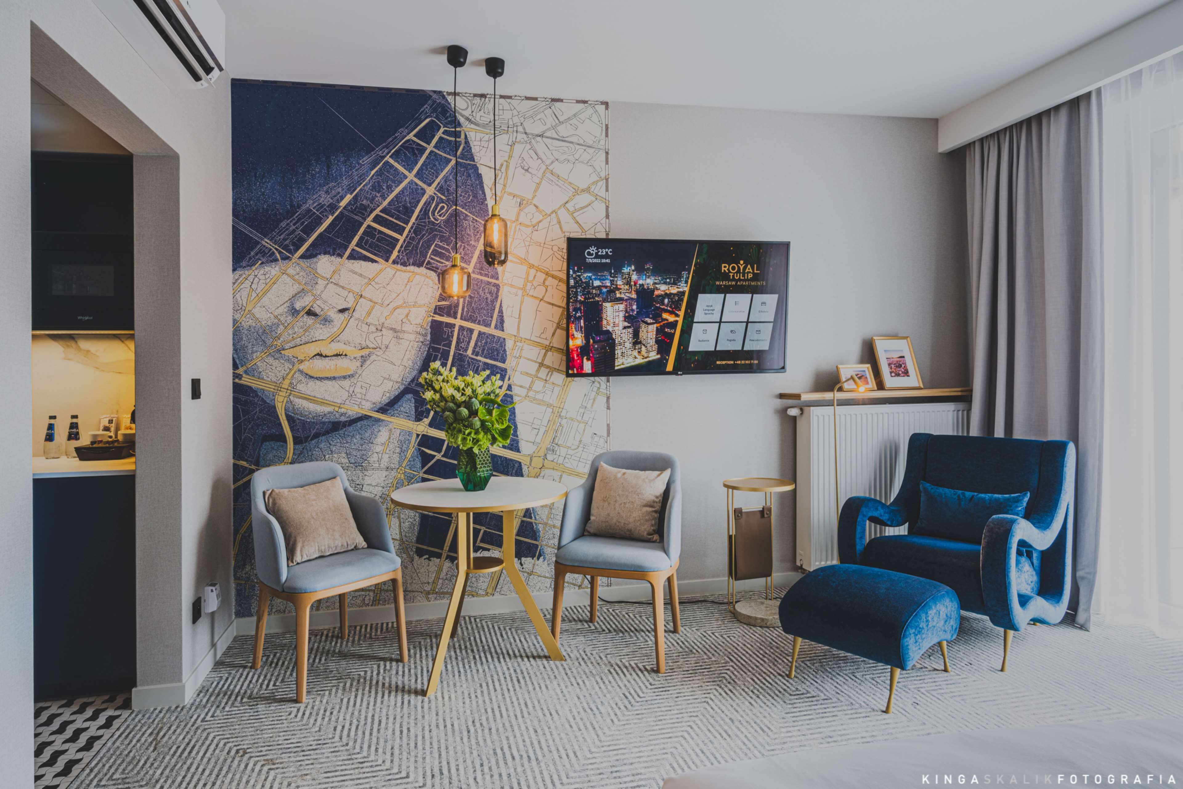 A modern hotel room with a large abstract painting, two armchairs, a round table with a vase, a blue armchair, and a TV displaying a cityscape.