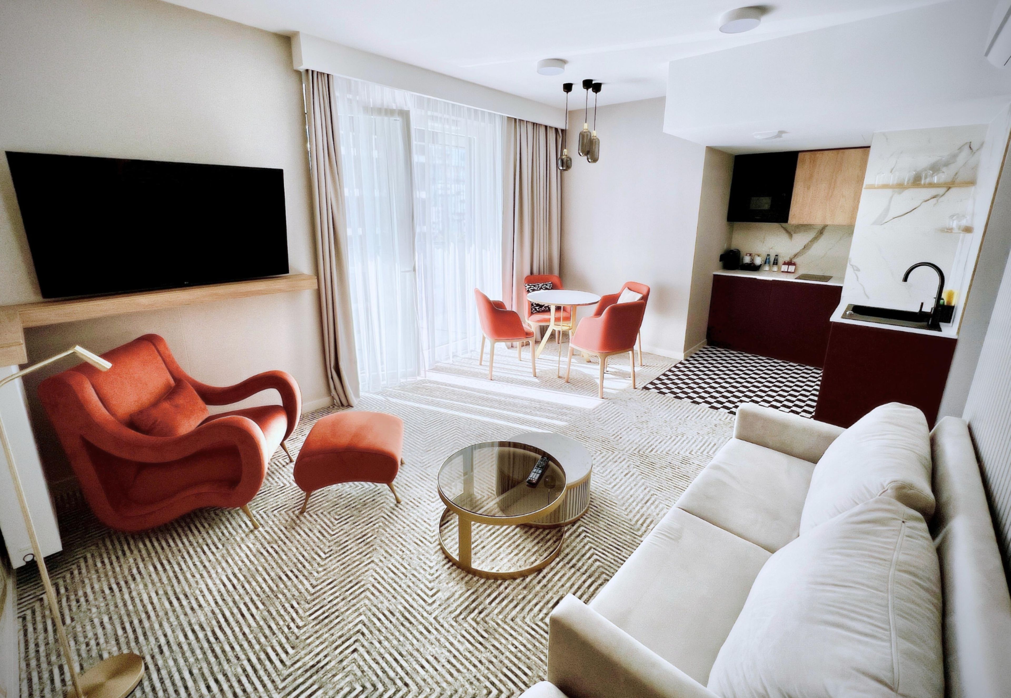 A modern hotel room with a flat-screen TV, a sofa, a dining area with chairs, and a kitchen area.