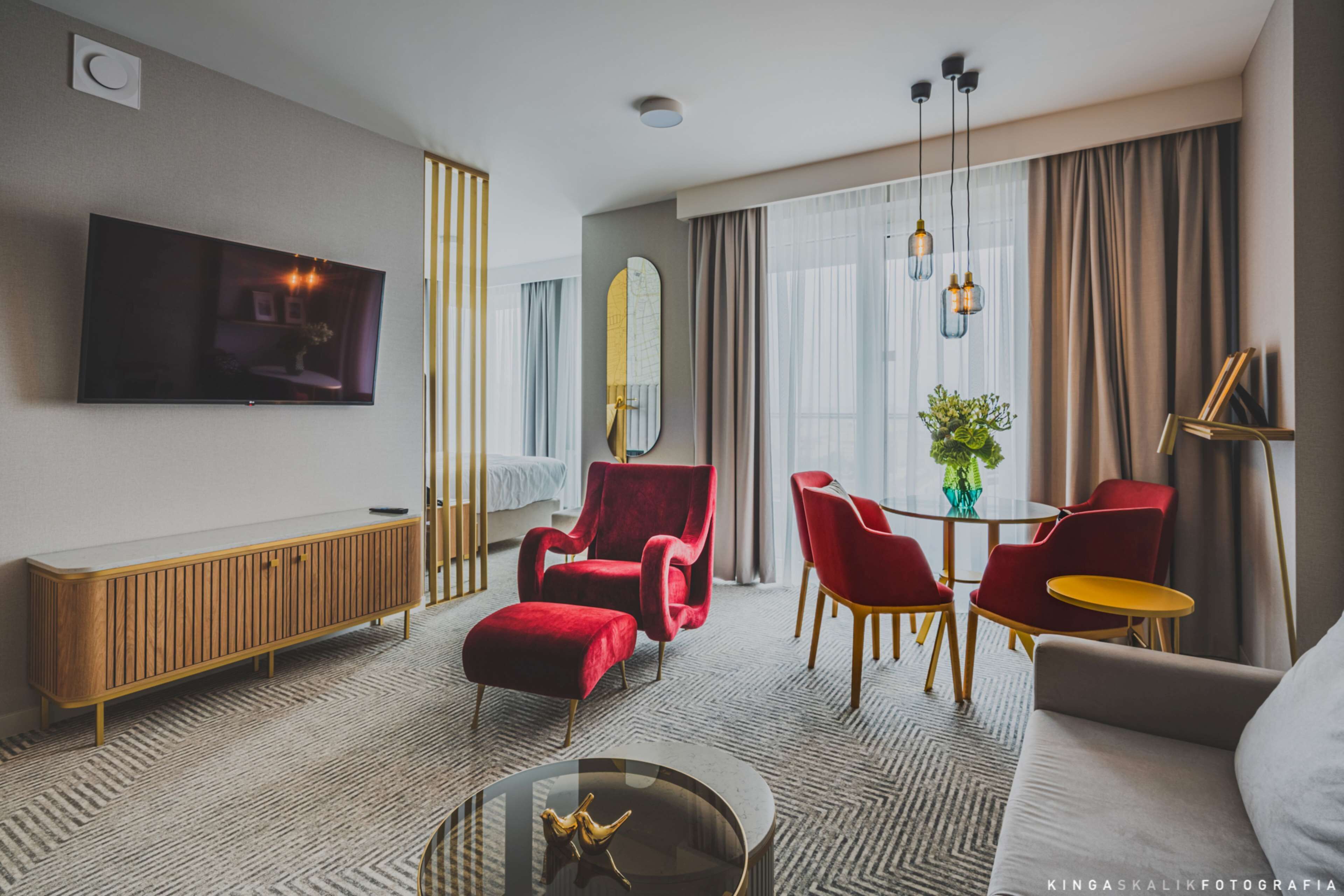 A modern hotel room with a TV, red chairs, a sofa, a dining table, and a large window with curtains.