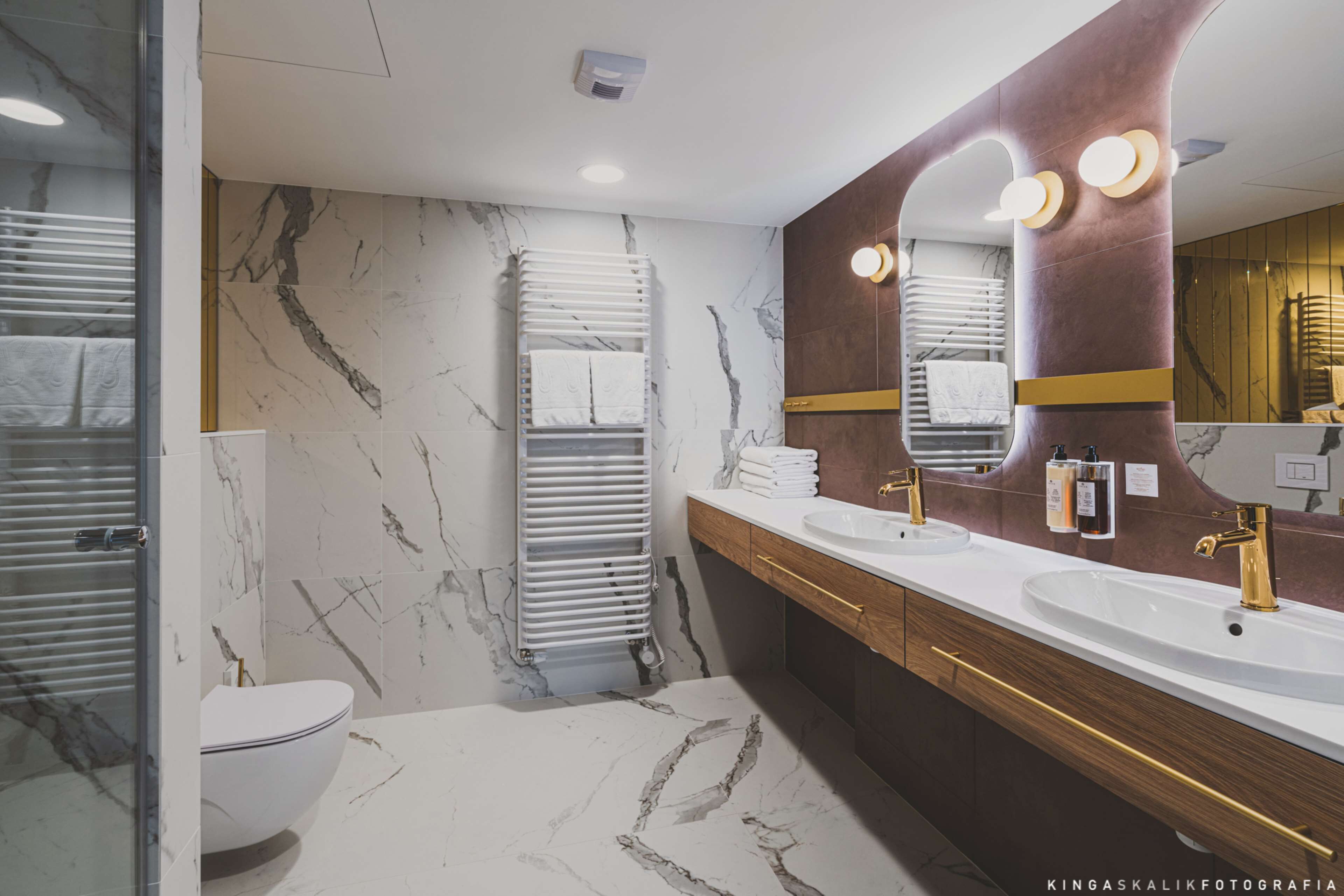 Een moderne badkamer met marmeren vloeren, twee wastafels met gouden kranen en een grote spiegel met verlichting.