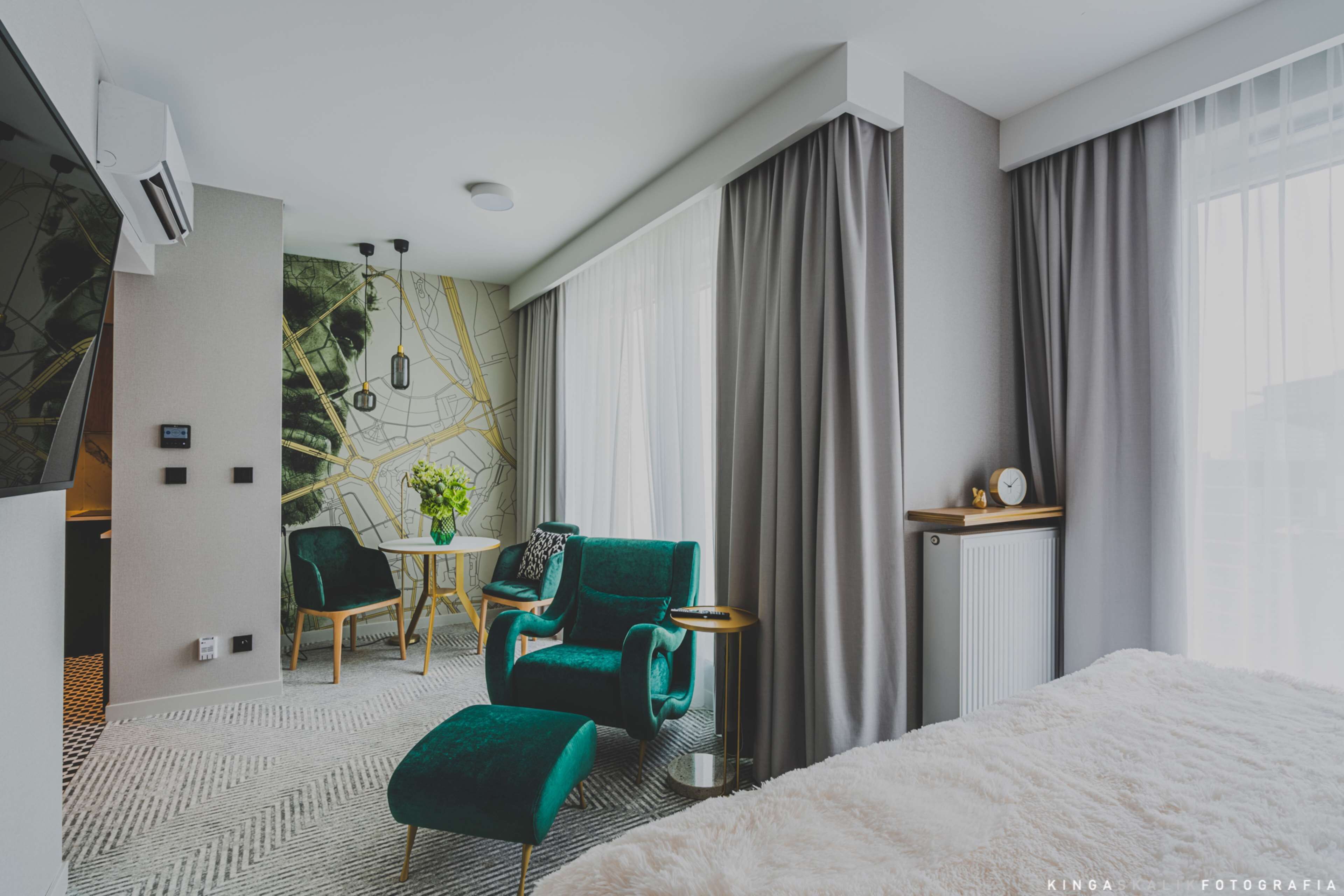 A modern hotel room with a large bed, a green armchair, a small round table, and a view of the city through large windows.