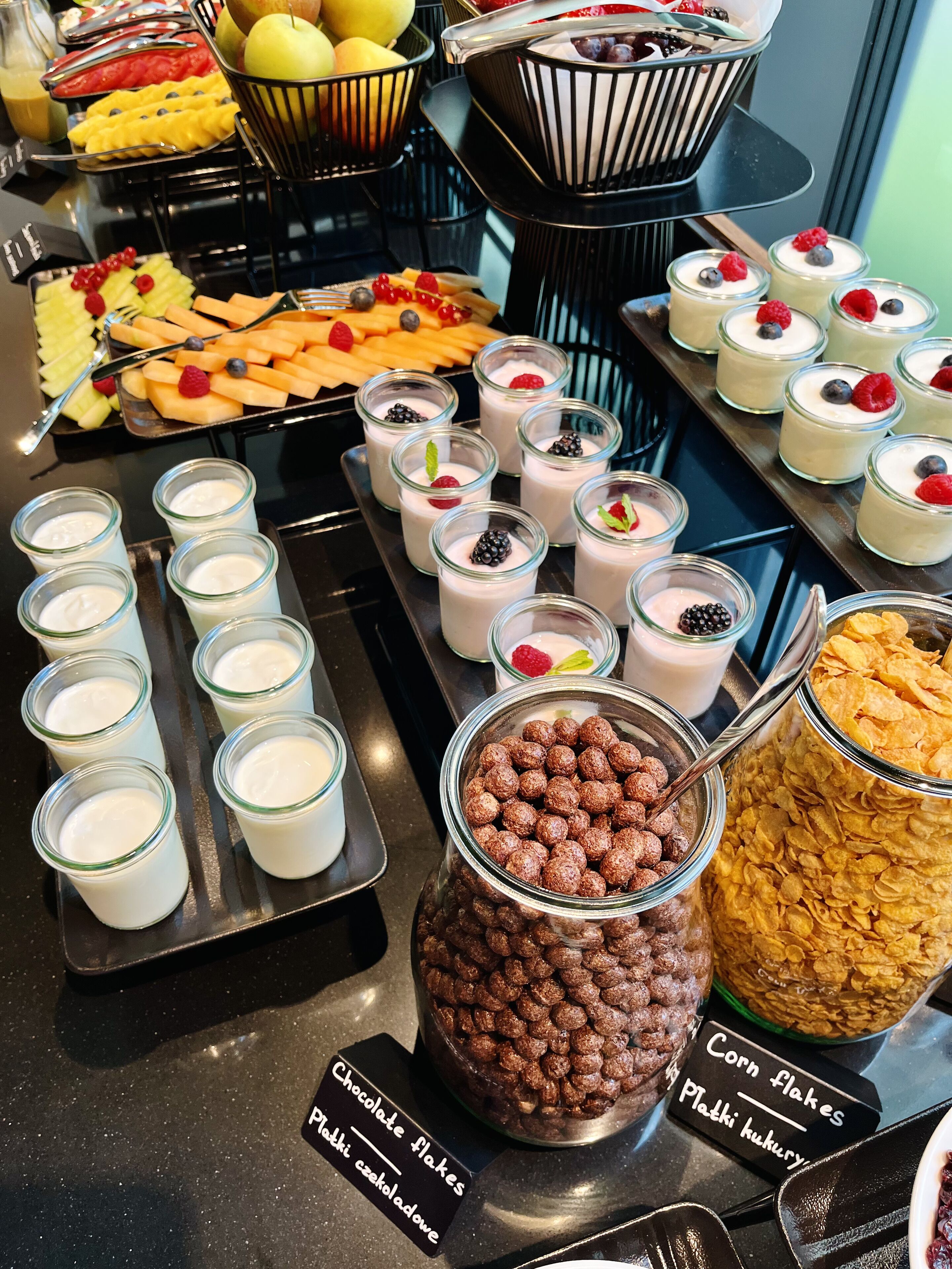 A buffet with various food items including chocolate flakes, corn flakes, and a variety of fruits and desserts.