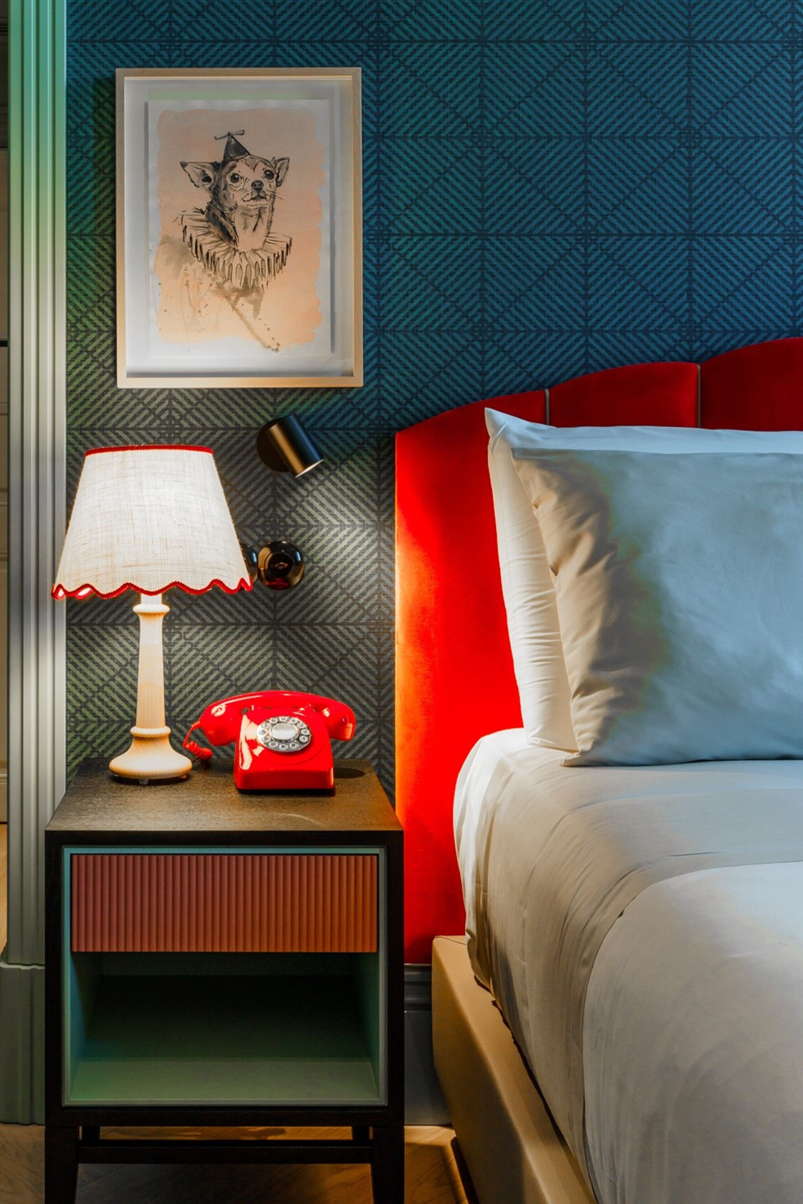 A neatly arranged bedroom with a red telephone, a framed picture on the wall, and a bedside lamp.
