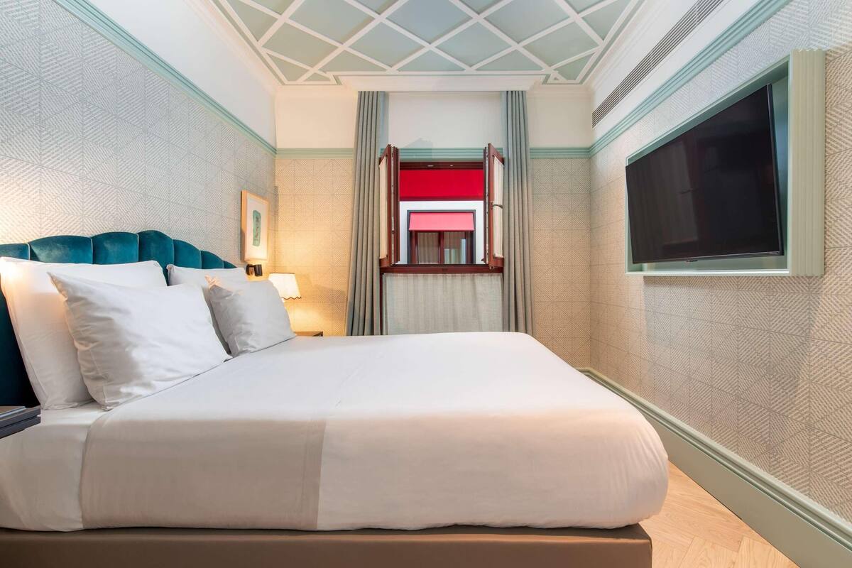 Habitación de hotel con cama, televisor montado en la pared, ventana y vista a una estructura roja y blanca en el exterior.