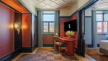 Et hotelværelse med et ternet gulv, et skrivebord med stol, et fjernsyn og et vindue med udsigt til bygninger.