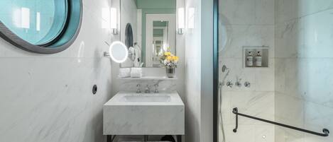 A modern bathroom with a marble sink, a round mirror, a handheld showerhead, and a glass-enclosed shower.