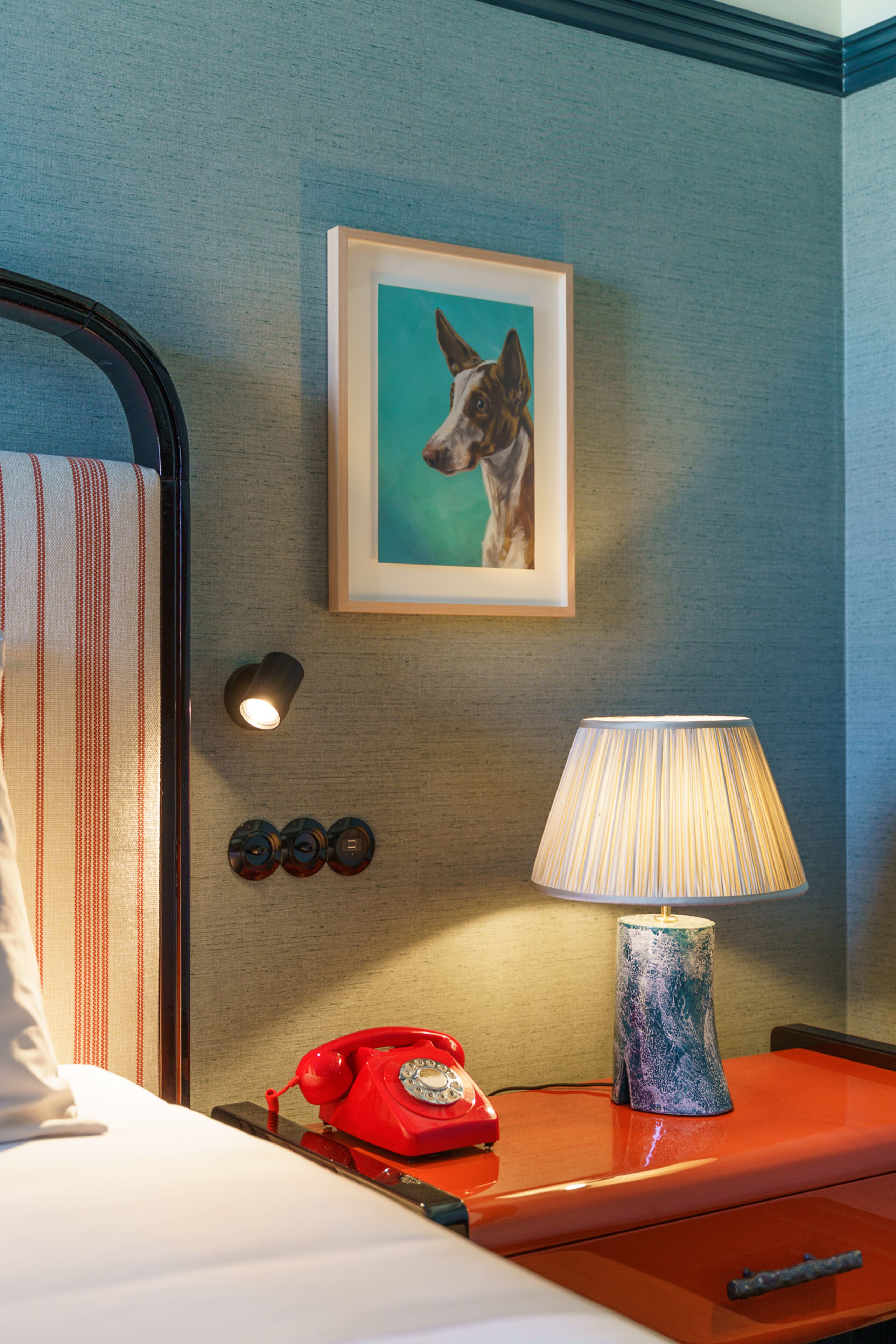 A hotel room with a bed, a red telephone, a lamp, a framed picture of a dog, and a wall-mounted light.