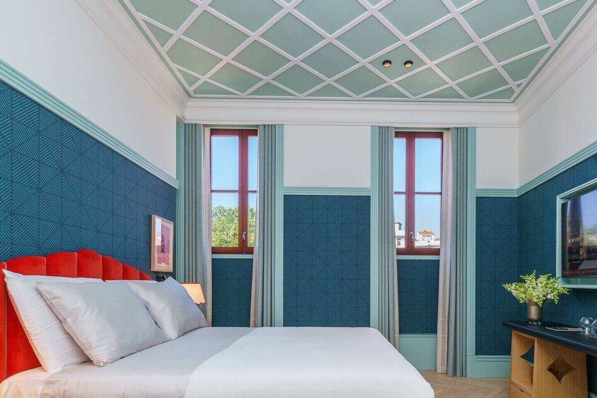 Un dormitorio con una cama, cabecera roja, dos ventanas, un televisor y una mesita con una planta.