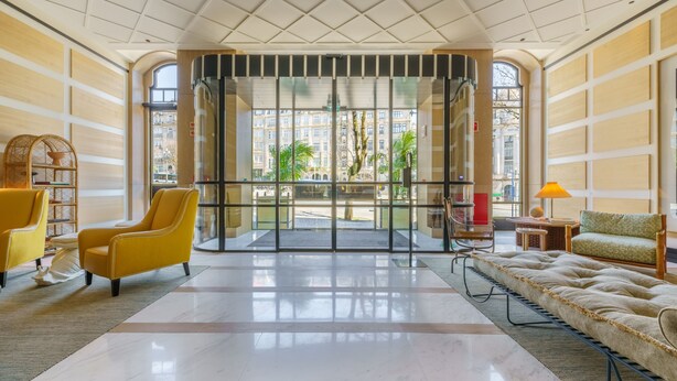 Un hall d’entrée spacieux avec une grande porte vitrée donnant sur l’extérieur, un fauteuil jaune, un canapé et une petite table avec une lampe.