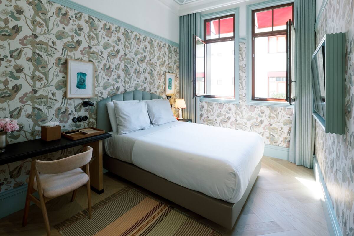 Un dormitorio con cama, escritorio con silla, un cuadro enmarco y ventana con cortinas.