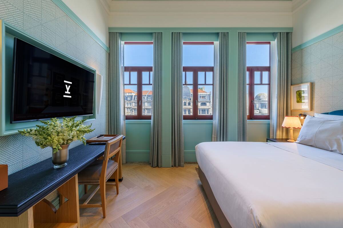 Habitación de hotel con cama, escritorio con televisor, lámpara y vista a edificios.