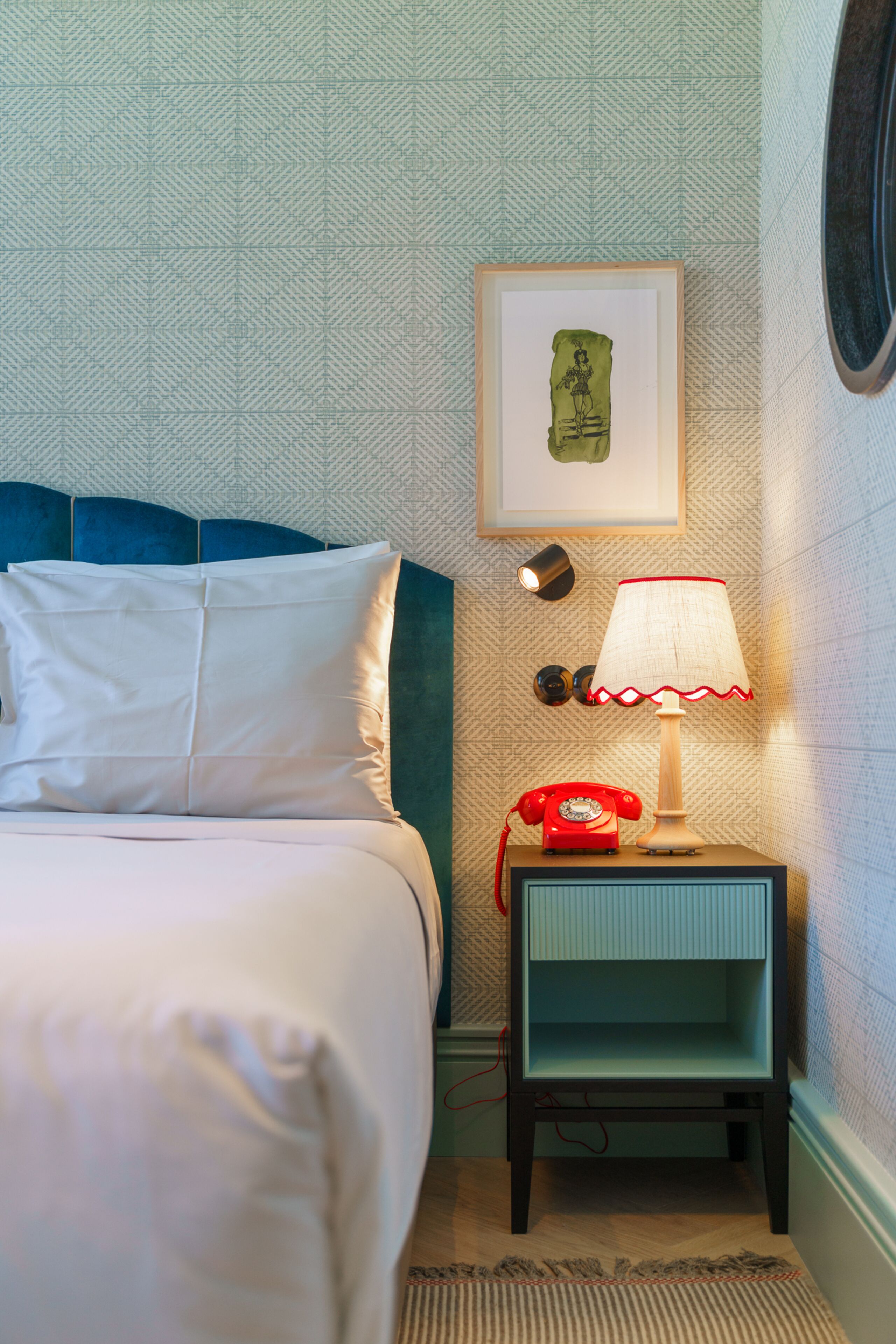 Een netjes ingerichte hotelkamer met een bed, een nachtkastje met een lamp, een lijst met een afbeelding aan de muur en een behang met patroon.
