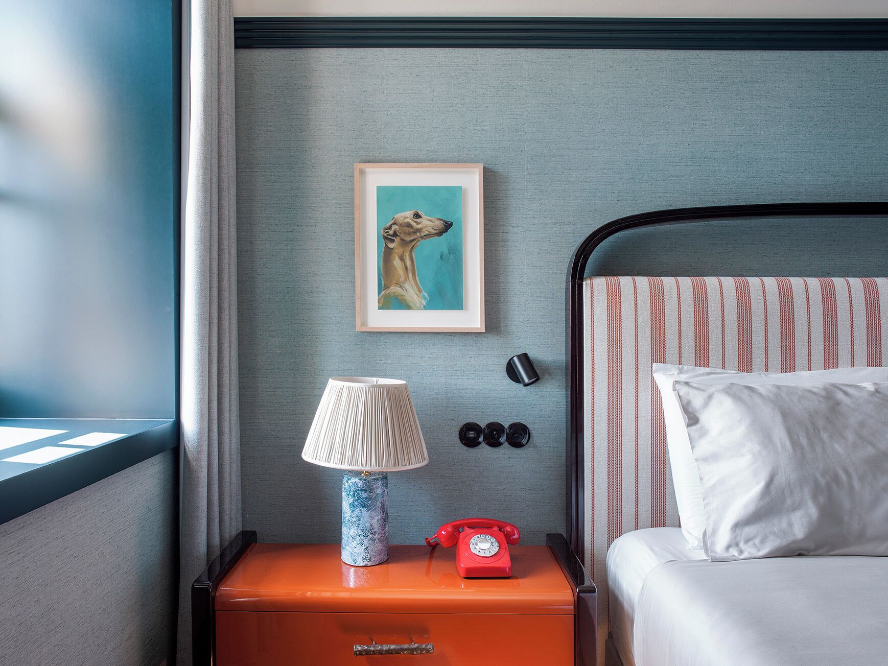Et hotelværelse med en seng, et natbord med en lampe, et indrammet billede og en rød telefon.