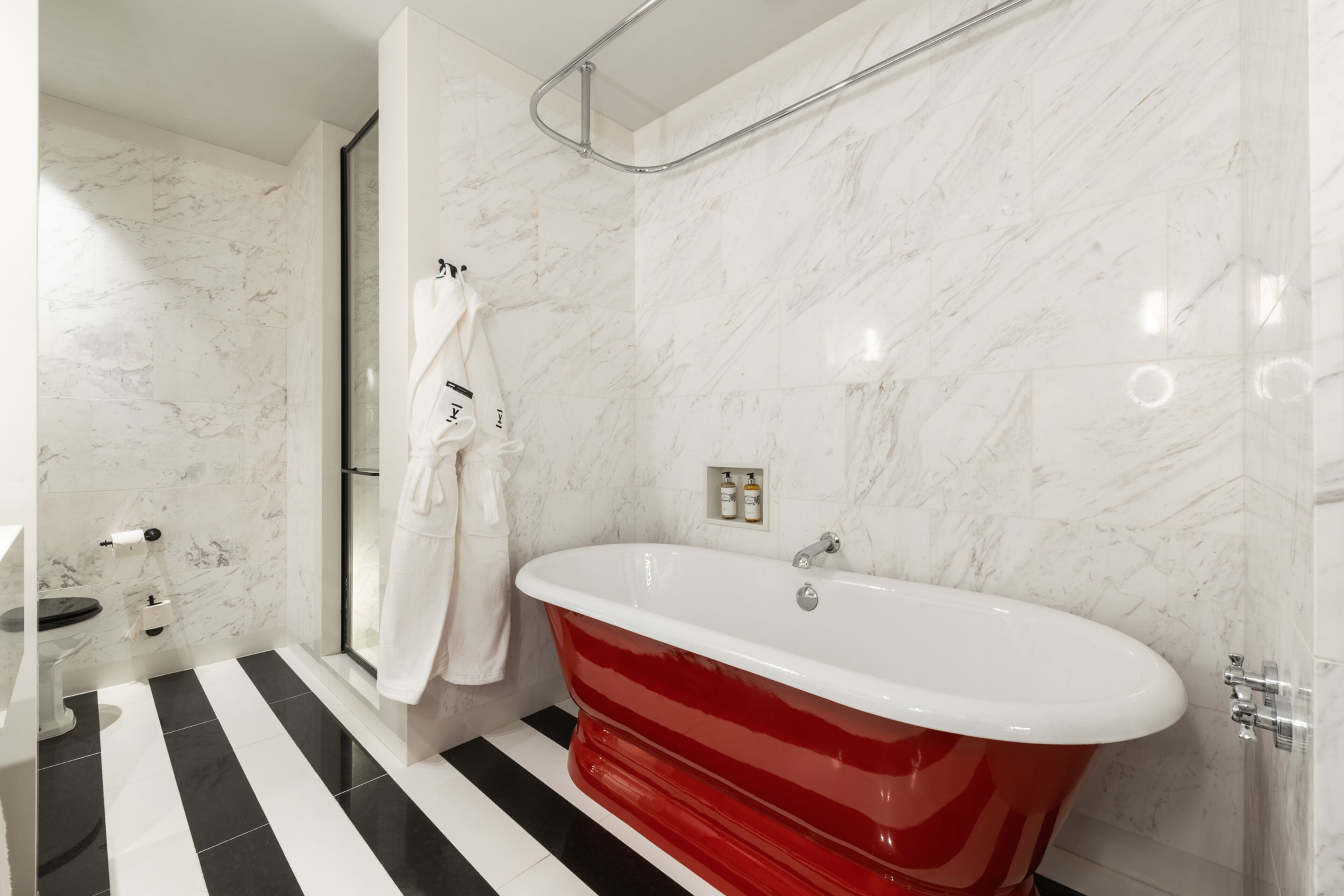 A modern bathroom with a red clawfoot bathtub, a glass shower enclosure, and a black and white striped floor.