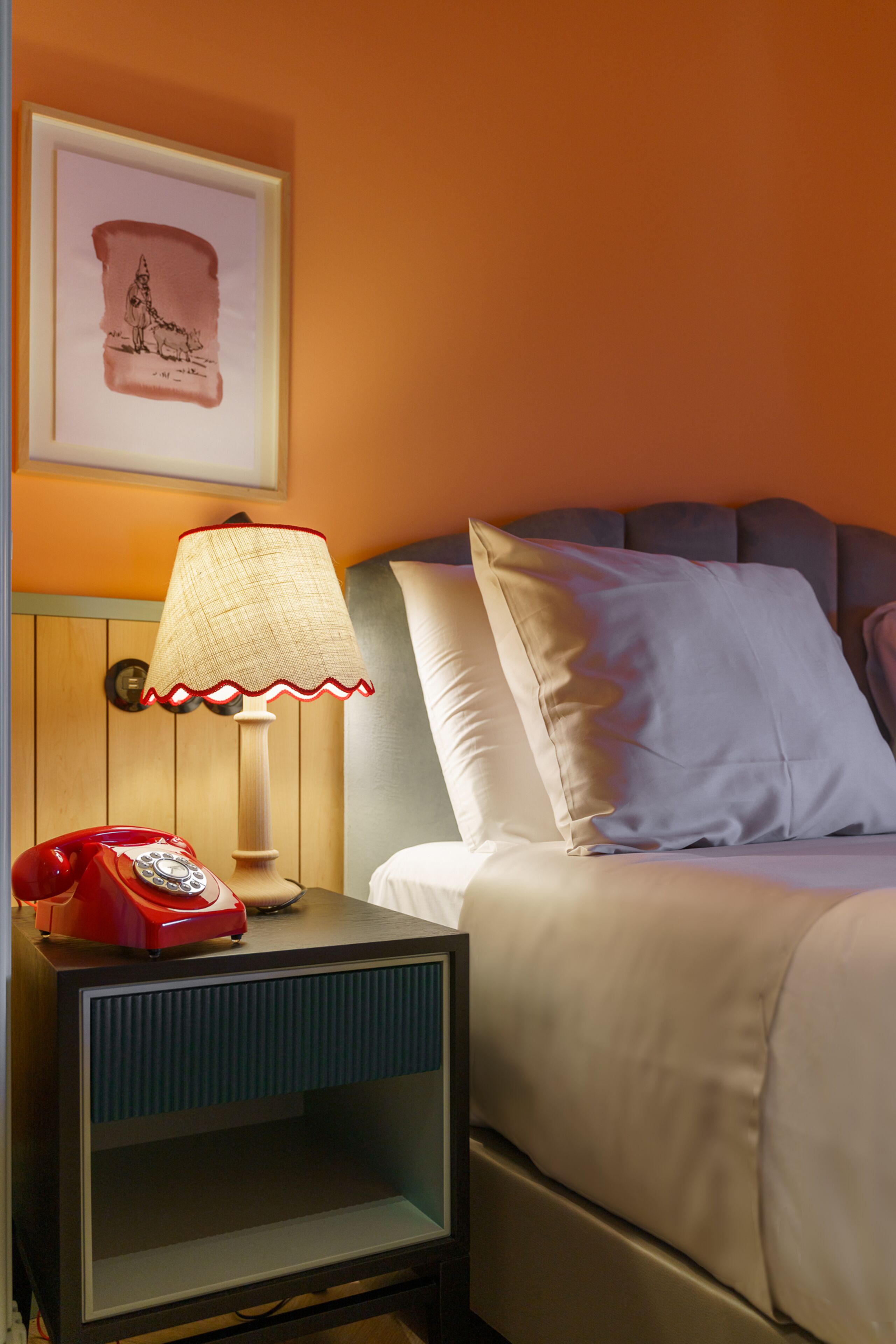 A neatly made bed with a headboard, a bedside lamp, and a vintage red telephone on a wooden nightstand.