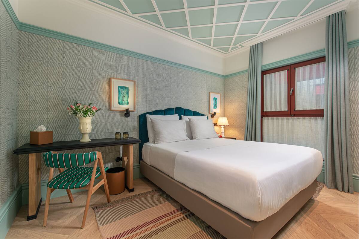 Habitación de hotel con cama, escritorio, silla, un florero con flores y cuadros en la pared.