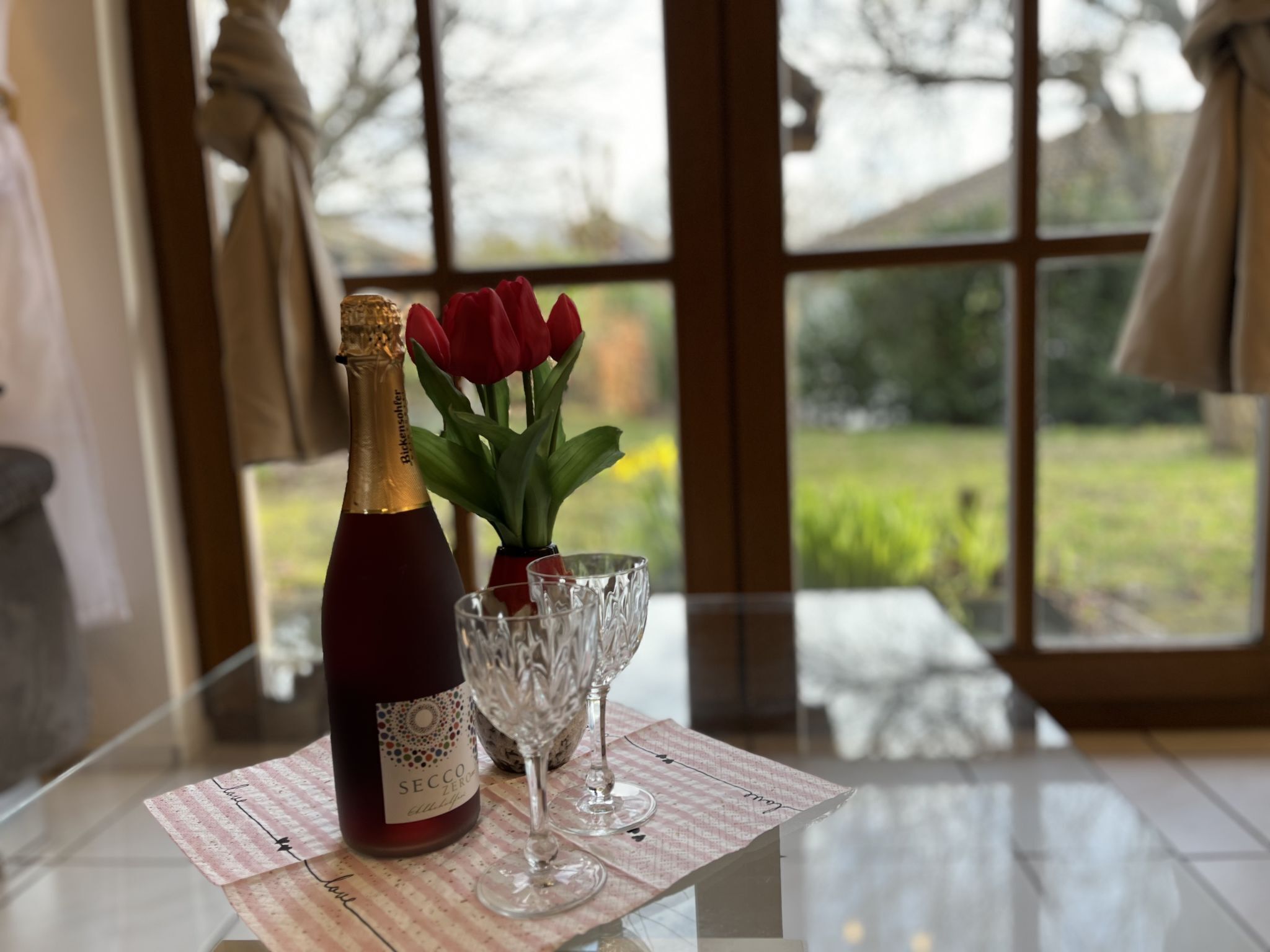 A bottle of Secco Zero wine, two wine glasses, and a vase with red tulips on a table with a checkered placemat.
