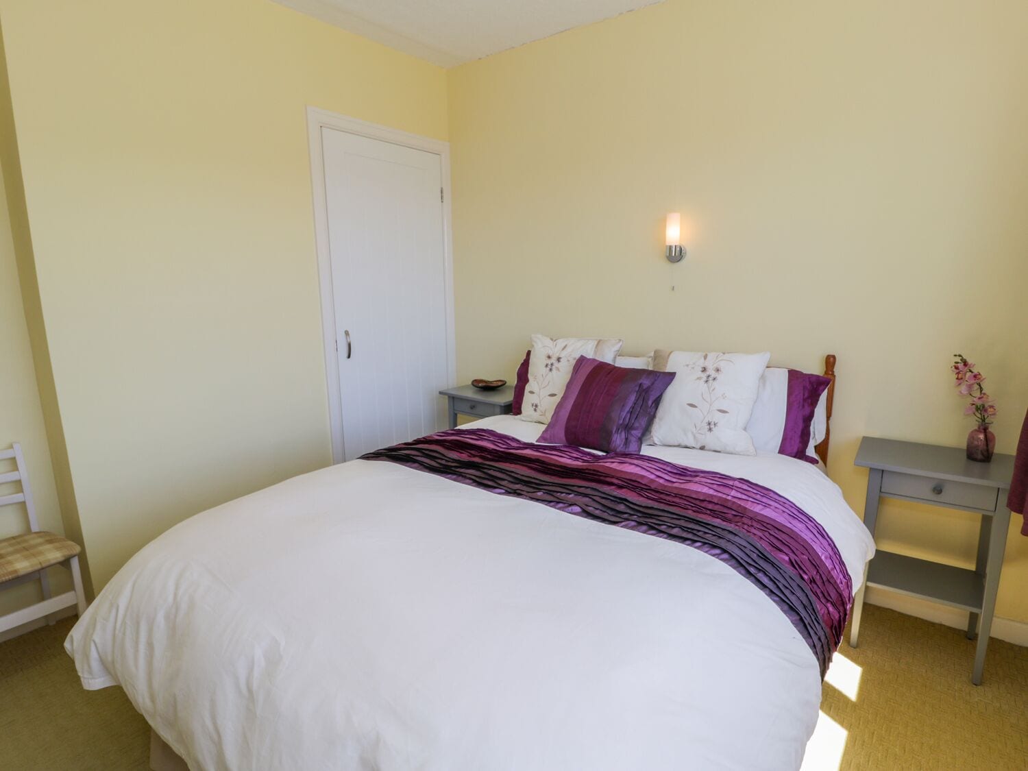 A bedroom with a bed, a bedside table, a nightstand, a chair, and a door.