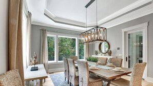 A dining area with a wooden table and wicker chairs, large windows, and a hanging light fixture. - Brand New Construction 3rd Row Home in Palmetto Dunes! (Hilton Head Island)