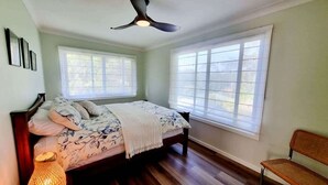 Une chambre à coucher avec un lit, un ventilateur de plafond, deux fenêtres, une chaise et une lampe.