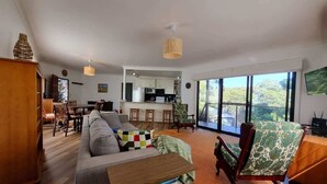Un vaste espace de vie comprenant un canapé, une table basse et un fauteuil. Au fond, on aperçoit une cuisine avec une grande fenêtre.