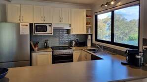 Une cuisine moderne avec des meubles blancs, un réfrigérateur en acier inoxydable, un four à micro-ondes, une cuisinière et une grande fenêtre donnant sur les arbres.