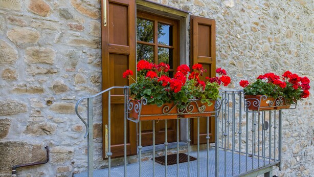 Un bâtiment en pierre avec des volets en bois, un balcon en métal orné de jardinières et une rambarde en métal.