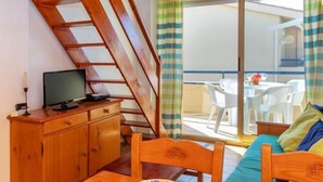 Une pièce avec un escalier en bois, un téléviseur posé sur un meuble en bois, une table à manger avec des chaises et un canapé agrémenté de coussins colorés.