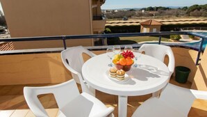 Une table ronde entourée de chaises blanches sur un balcon, un saladier rempli de fruits et des verres d’eau.