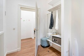 Un pasillo con una puerta blanca, un radiador, un reposapiés azul y un estante blanco con una chaqueta y una lámpara.
