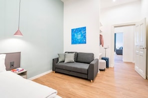 Una habitación de hotel moderna con un sofá gris, una mesita de noche pequeña con libros, un cuadro abstracto azul en la pared y un suelo de madera.