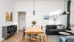 A modern living room with a wooden dining table, a black sofa, and a grey sofa. There is a bookshelf on the left and a small table with a vase of flowers on the right.