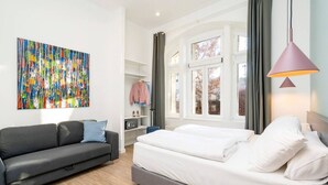 A modern hotel room with a grey sofa, a bed with white linens, a colorful abstract painting on the wall, and a large window with curtains.