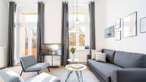 A modern living room with a grey sofa, two armchairs, a coffee table, and a TV. There are large windows with curtains, a hanging light, and framed pictures on the wall.